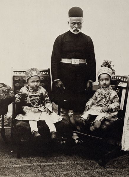 Junge Prinzessinnen mit Aufseher. by Raja Lala Deen Dayal. 1900. Motifs recorded: kinder, mann, teppich, foto, gürtel, stuhl, sitzen, bart.